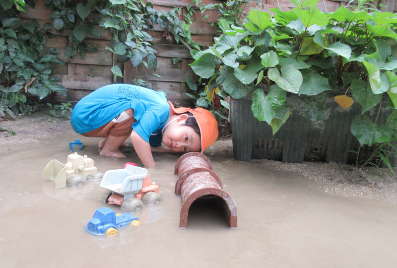 水たまりの中でおもちゃのトンネルを覗く園児